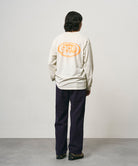 Back view of a model wearing a natural pigment colored Gramicci GCE Tee Pigment Dyed with a graphic and black pants on a light-tone background.