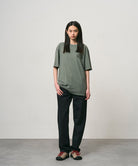 Model wearing a sage pigment colored Gramicci GCE Tee Pigment Dyed and black pants on a light-tone background.