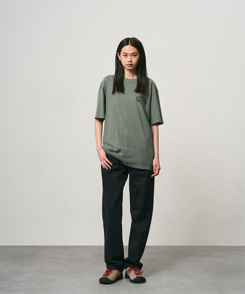 Model wearing a sage pigment colored Gramicci GCE Tee Pigment Dyed and black pants on a light-tone background.