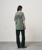 Back view of a model wearing a sage pigment colored Gramicci GCE Tee Pigment Dyed with a graphic and black pants on a light-tone background.