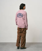 Back view of a model wearing a lilac pigment Gramicci GCE Long Sleeve Tee Pigment Dyed with a graphic and brown pants on a light-tone background.