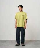 Model wearing a sweet pea colored Gramicci Earth Tee with a graphic and black pants on a light-tone background.