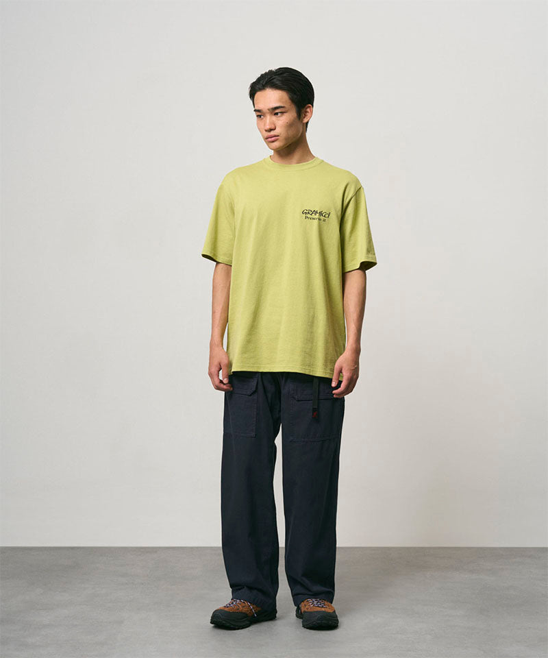 Model wearing a sweet pea colored Gramicci Earth Tee with a graphic and black pants on a light-tone background.