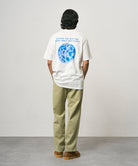 Back view of a model wearing a white Gramicci Earth Tee with a blue graphic and light olive pants on a light-tone background.