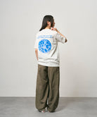 Back view of a model wearing a white Gramicci Earth Tee with a blue graphic and olive pants on a light-tone background.
