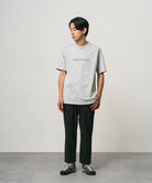 Model wearing a mist colored Gramicci Vase Tee and black pants on a light-tone background.