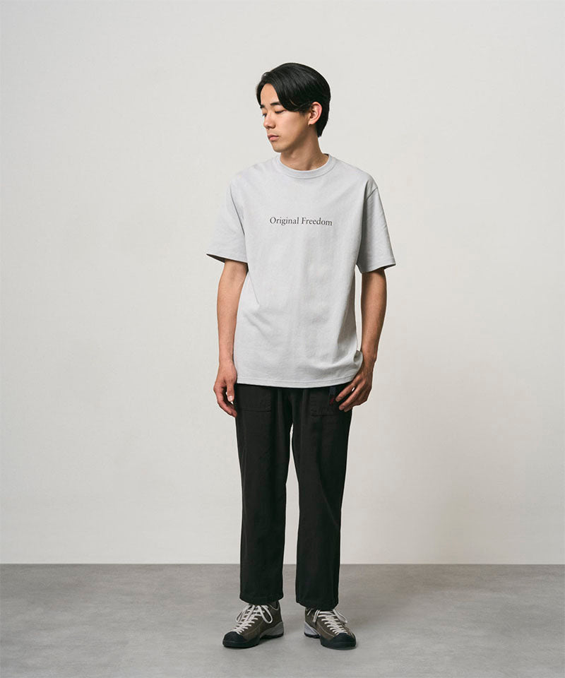 Model wearing a mist colored Gramicci Vase Tee and black pants on a light-tone background.