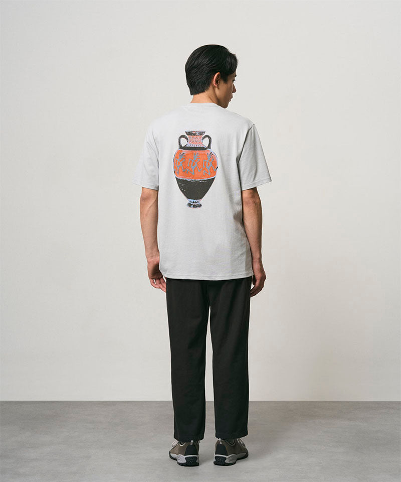 Back view of a model wearing a mist colored Gramicci Vase Tee with a graphic and black pants on a light-tone background.