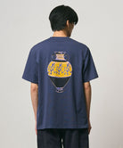 Back view of a model wearing a Gramicci navy Vase Tee with a graphic against a light-tone background.