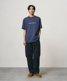 Model wearing a navy Gramicci Vase Tee and black pants on a light-tone background.