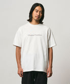 Model wearing a Gramicci white Vase Tee with a graphic against a light-tone background.