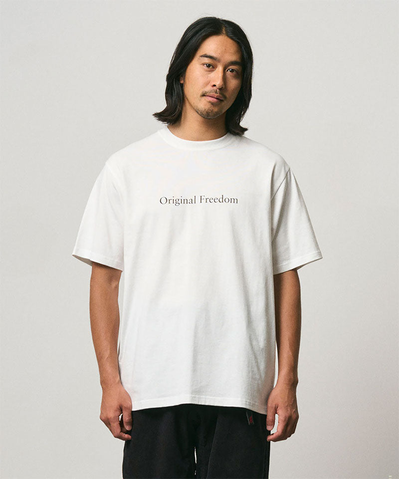 Model wearing a Gramicci white Vase Tee with a graphic against a light-tone background.
