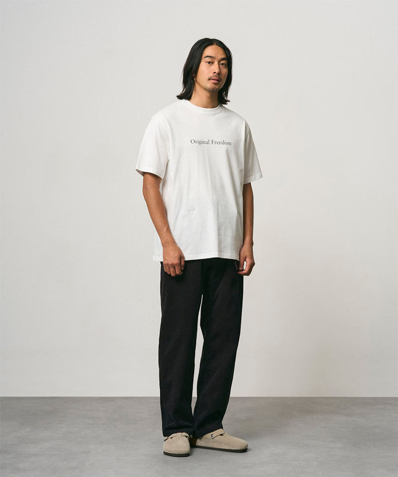 Model wearing a white Gramicci Vase Tee and black pants on a light-tone background.