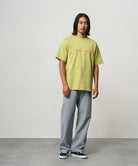 Model wearing a sweet pea colored Gramicci Starburst Tee with a graphic and grey pants on a light-tone background.