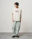 Model wearing a natural pigment colored Gramicci Bone Tee Pigment Dyed with a graphic and light green pants on a light-tone background.