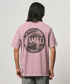 Back view of a model wearing a Gramicci lilac pigment colored Icon Tee Pigment Dyed with a graphic against a light-tone background.