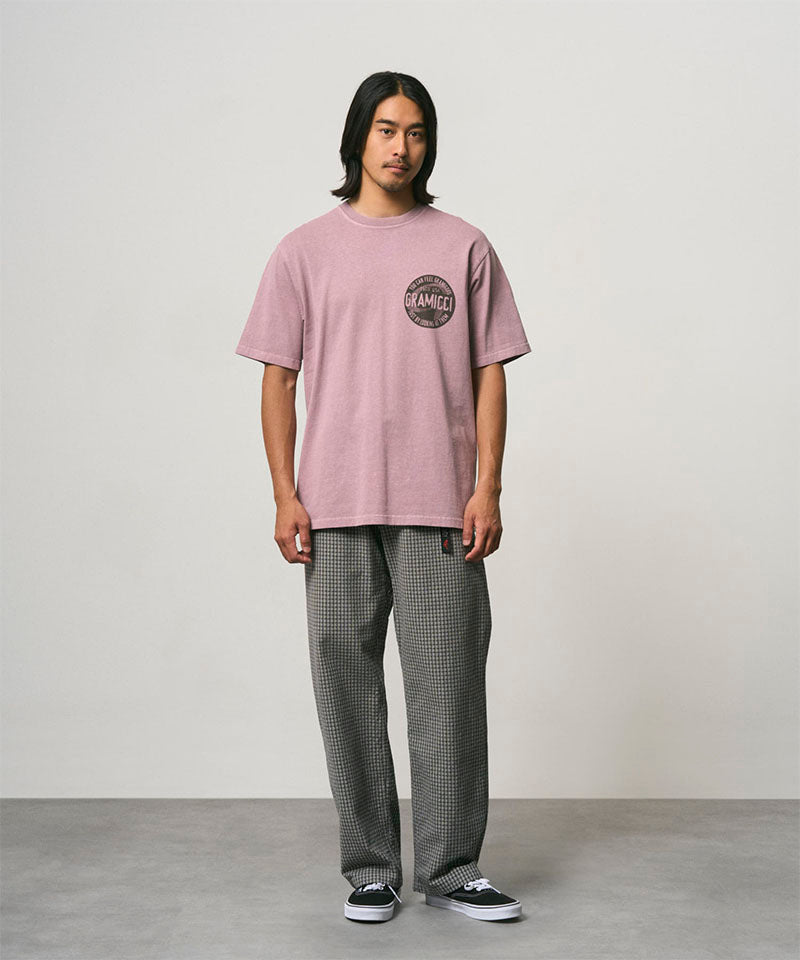 Model wearing a lilac pigment colored Gramicci Icon Tee Pigment Dyed with a graphic and grey pants on a light-tone background.