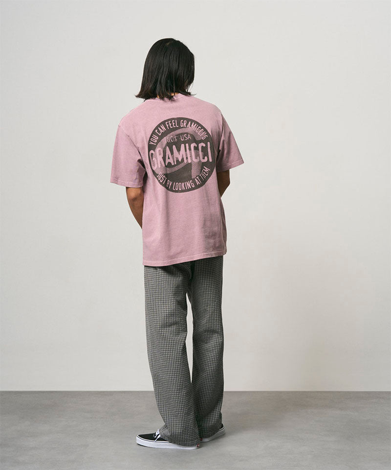 Back view of a model wearing a lilac pigment colored Gramicci Icon Tee Pigment Dyed with a graphic and grey pants on a light-tone background.