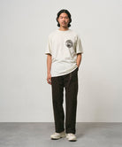 Model wearing a natural pigment colored Gramicci Icon Tee Pigment Dyed with a graphic and dark brown pants on a light-tone background.
