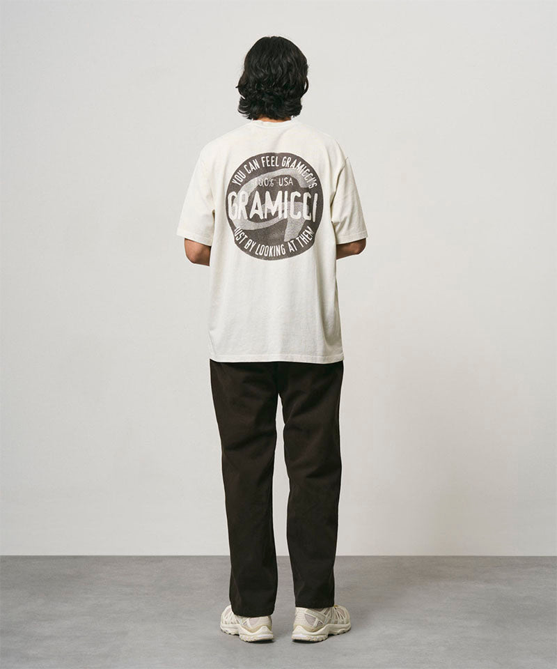 Back view of a model wearing a natural pigment colored Gramicci Icon Tee Pigment Dyed with a graphic and dark brown pants on a light-tone background.
