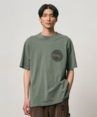 Model wearing a Gramicci sage pigment colored Icon Tee Pigment Dyed with a graphic against a light-tone background.