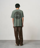 Back view of a model wearing a sage pigment colored Gramicci Icon Tee Pigment Dyed with a graphic and dark brown pants on a light-tone background.