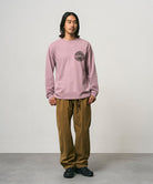 Model wearing a lilac pigment colored Gramicci Icon Long Sleeve Tee Pigment Dyed with a graphic and olive brown pants on a light-tone background.