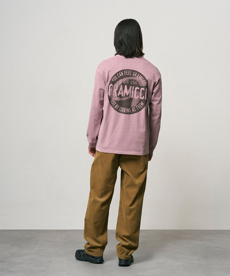 Back view of a model wearing a lilac pigment colored Gramicci Icon Long Sleeve Tee Pigment Dyed with a graphic and olive brown pants on a light-tone background.