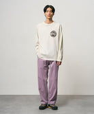 Model wearing a natural pigment colored Gramicci Icon Long Sleeve Tee Pigment Dyed with a graphic and lavender pants on a light-tone background.