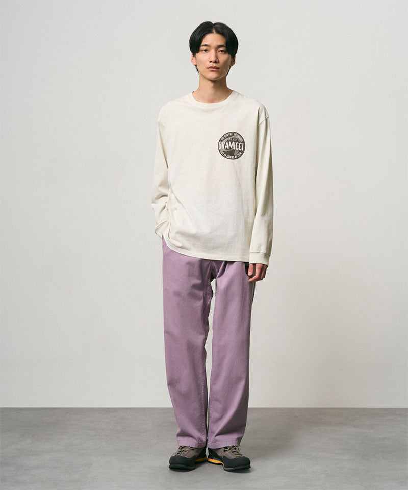Model wearing a natural pigment colored Gramicci Icon Long Sleeve Tee Pigment Dyed with a graphic and lavender pants on a light-tone background.