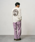 Back view of a model wearing a natural pigment colored Gramicci Icon Long Sleeve Tee Pigment Dyed with a graphic and lavender pants on a light-tone background.
