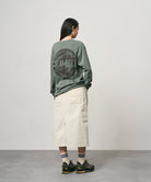 Back view of a model wearing a sage pigment colored Gramicci Icon Long Sleeve Tee Pigment Dyed with a graphic and creme colored skirt on a light-tone background.