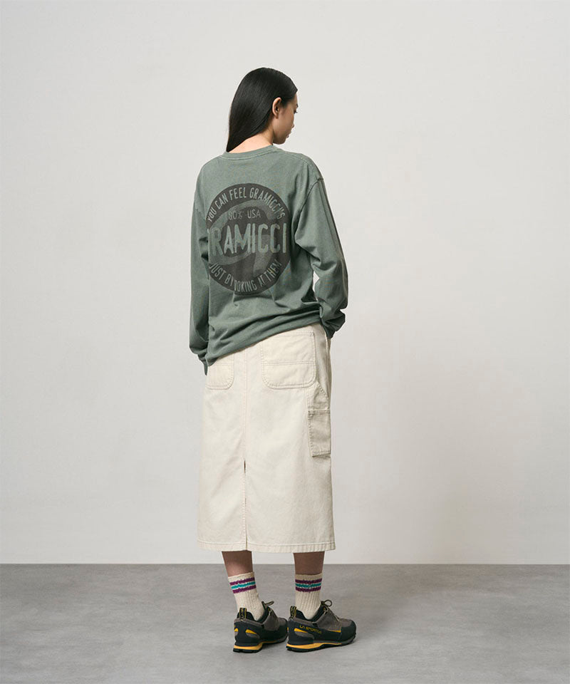 Back view of a model wearing a sage pigment colored Gramicci Icon Long Sleeve Tee Pigment Dyed with a graphic and creme colored skirt on a light-tone background.