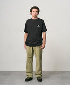 Model wearing a black Gramicci One Point Logo Tee with a graphic and light olive pants on a light-tone background.