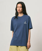 Model wearing a Gramicci dusky blue One Point Logo Tee with a graphic against a light-tone background.