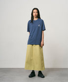 Model wearing a dusky blue Gramicci One Point Logo Tee with a graphic and light olive skirt on a light-tone background.