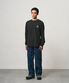 Model wearing a black Gramicci One Point Logo Long Sleeve Tee with a graphic and blue pants on a light-tone background.