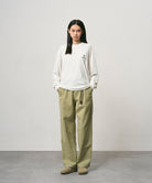 Model wearing a white Gramicci One Point Logo Long Sleeve Tee with a graphic and light olive pants on a light-tone background.