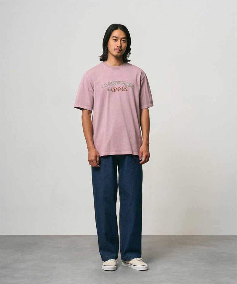Model wearing a lillac pigment colored Gramicci Livin' Tee Pigment Dyed with a Livin' Under A Rock graphic and blue pants on a light-tone background.