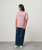 Model wearing a lillac pigment colored Gramicci Livin' Tee Pigment Dyed with a Livin' Under A Rock graphic and blue pants on a light-tone background.