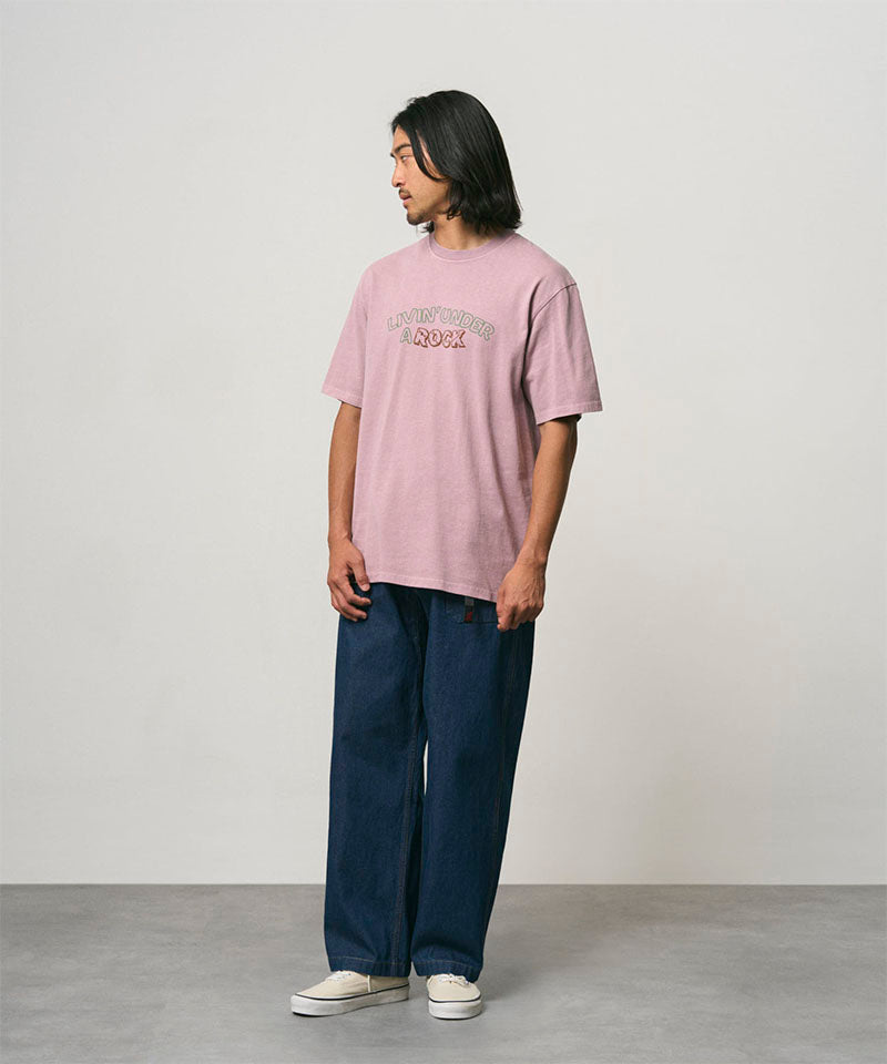 Model wearing a lillac pigment colored Gramicci Livin' Tee Pigment Dyed with a Livin' Under A Rock graphic and blue pants on a light-tone background.