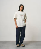 Model wearing a natural pigment colored Gramicci Livin' Tee Pigment Dyed with a Livin' Under A Rock graphic and blue pants on a light-tone background.