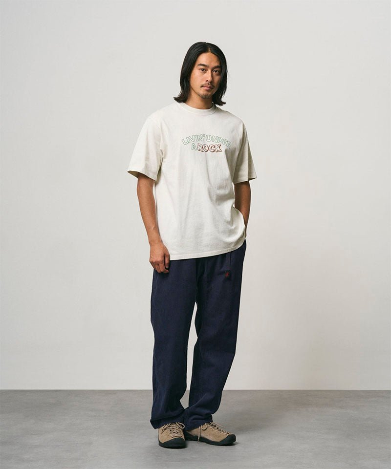 Model wearing a natural pigment colored Gramicci Livin' Tee Pigment Dyed with a Livin' Under A Rock graphic and blue pants on a light-tone background.