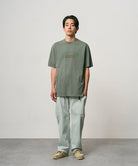 Model wearing a sage pigment colored Gramicci Livin' Tee Pigment Dyed with a Livin' Under A Rock graphic and light green pants on a light-tone background.