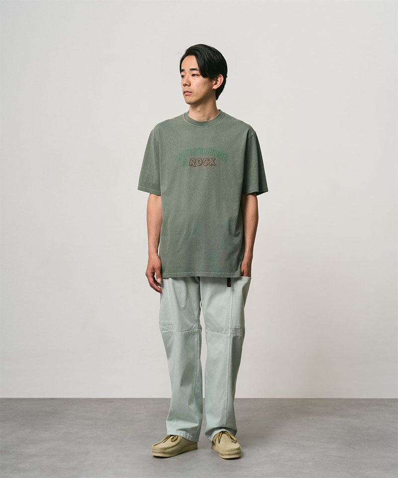 Model wearing a sage pigment colored Gramicci Livin' Tee Pigment Dyed with a Livin' Under A Rock graphic and light green pants on a light-tone background.