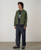 Model wearing a moss green Gramicci Taos Canvas Jacket and blue jeans on a light-tone background.
