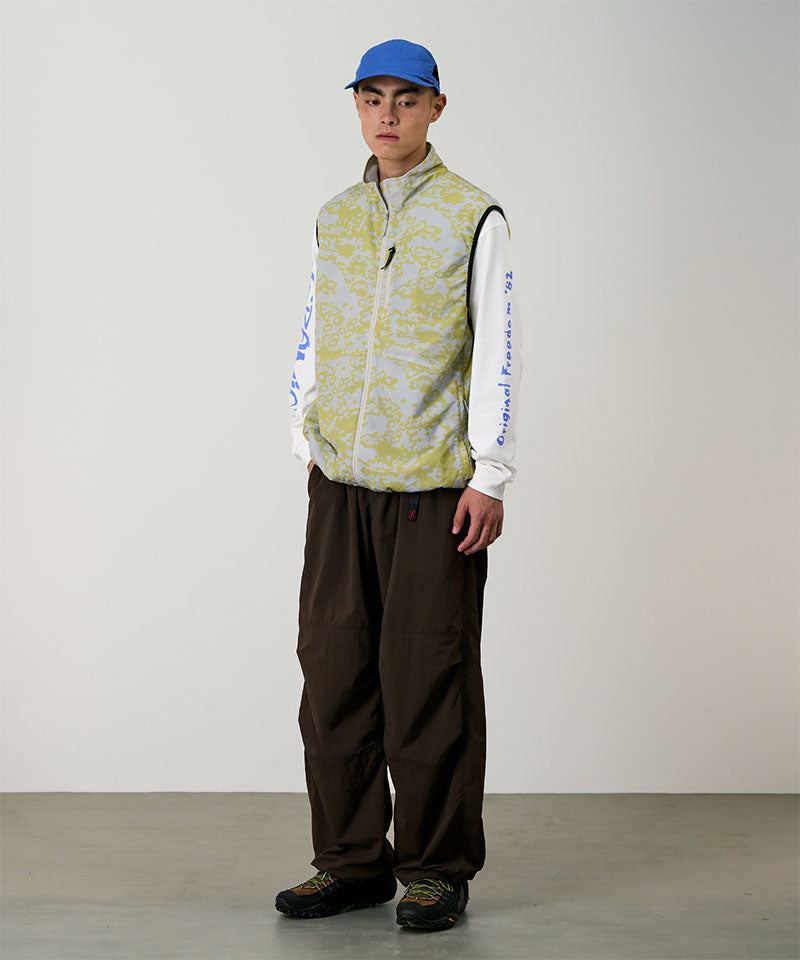 Person wearing a patterned vest, white long-sleeve shirt with text, brown pants, and blue cap against a plain background