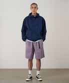 Model wearing a Gramicci Natural Dyed Jam Short in the color purple and a blue hoodie on a light-tone background.
