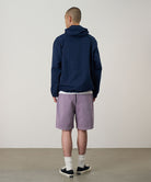 Back view of a model wearing a Gramicci Natural Dyed Jam Short in the color purple and a blue hoodie on a light-tone background.
