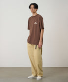 Model wearing a Natural Dyed Gramicci Pant Straight Fit in chino and brown shirt on a light-tone background.
